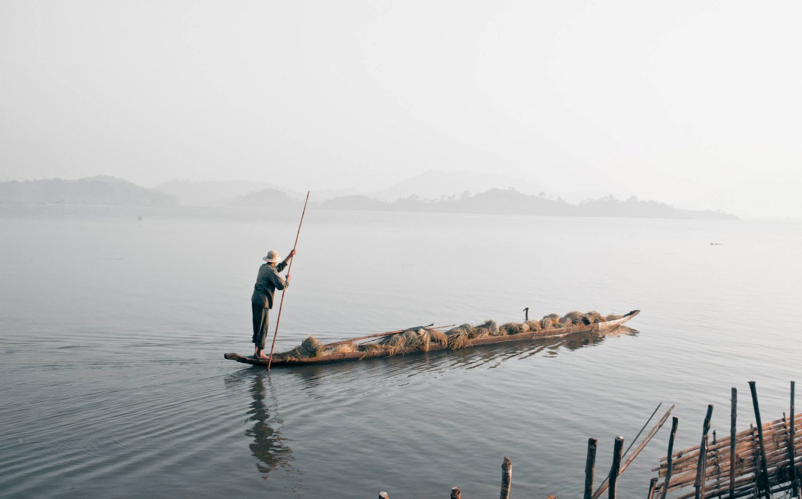 A man poling a wooden boat loaded with reeds across a misty lake near Buôn Ma Thuột