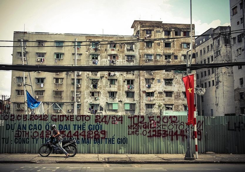 A weathered apartment block on a Chợ Lớn street, a motorbike passing along the corrugated fence out front