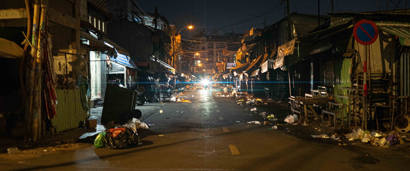 A dim alley market street at night in Ho Chi Minh City, litter scattered along the road, a motorbike headlight glowing in the distance