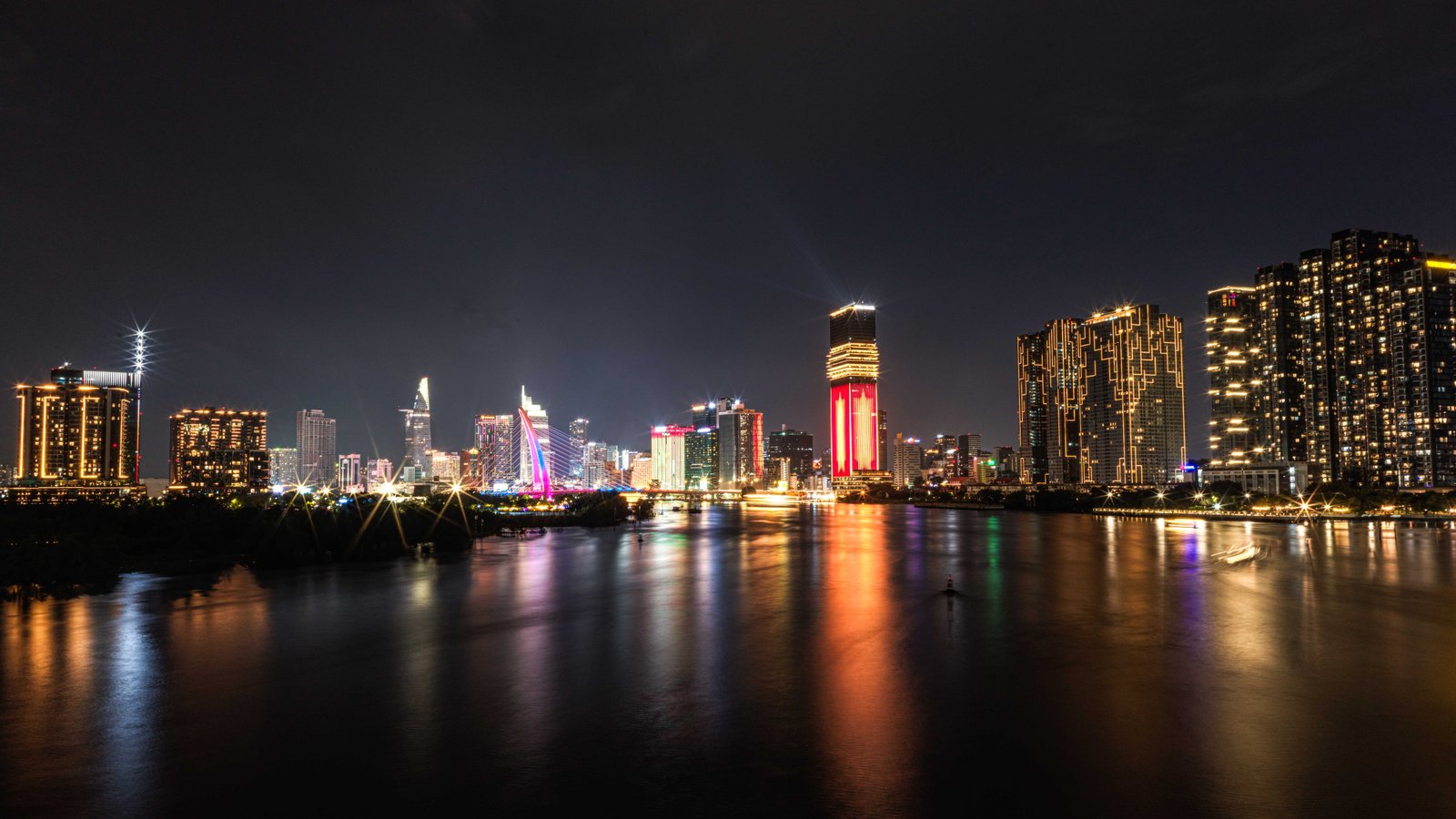 A night view of Ho Chi Minh City's skyline across the river, colorful lights reflecting on the water, boats passing below the bridge