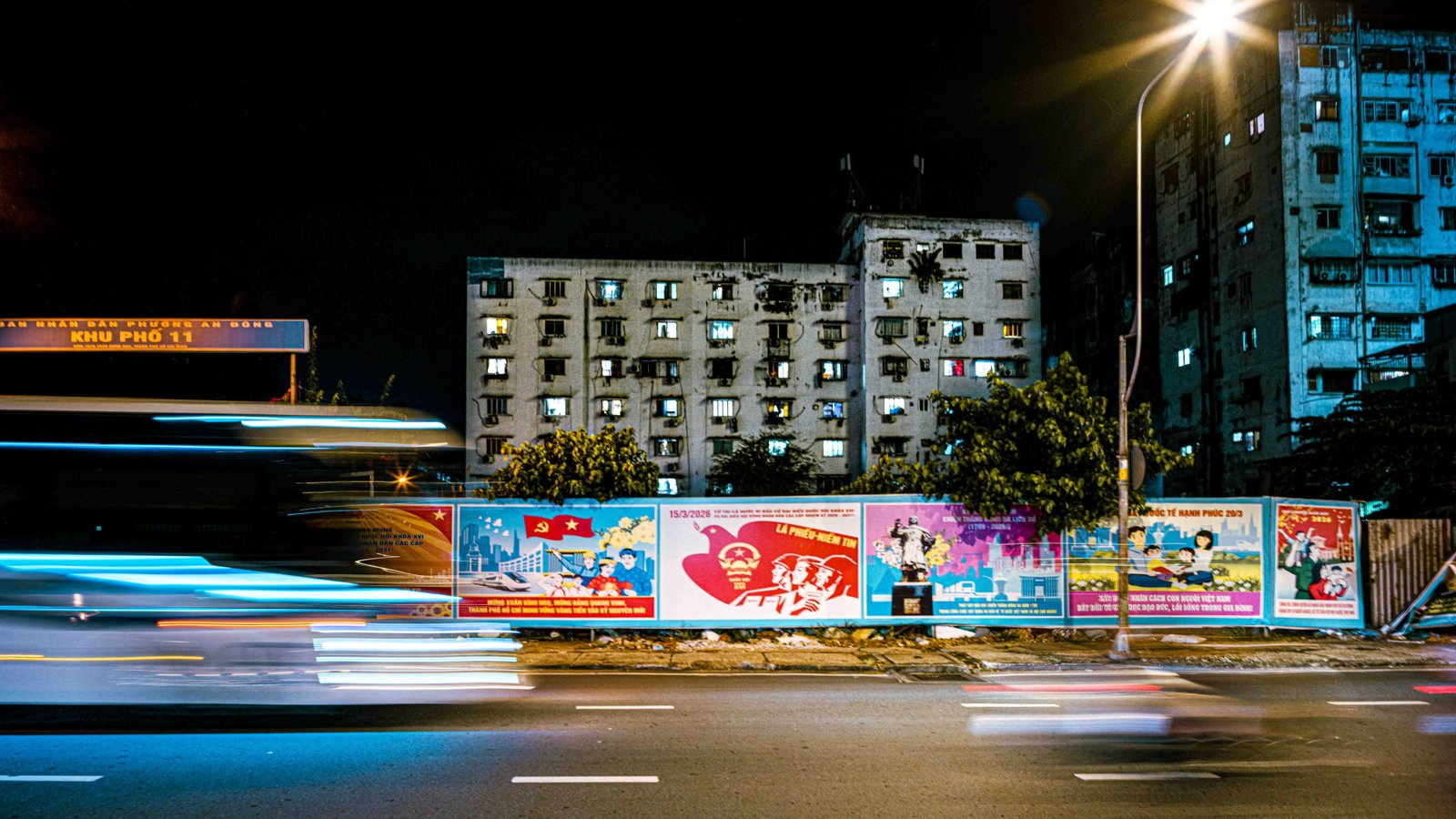 A long-exposure night shot of a bus streaking past propaganda murals and an aging apartment block in Ho Chi Minh City
