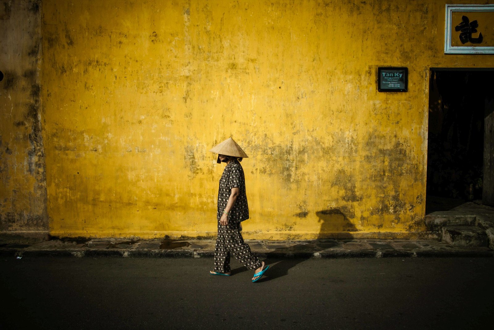 A woman in a conical hat walking past a weathered yellow wall in Hội An's old town