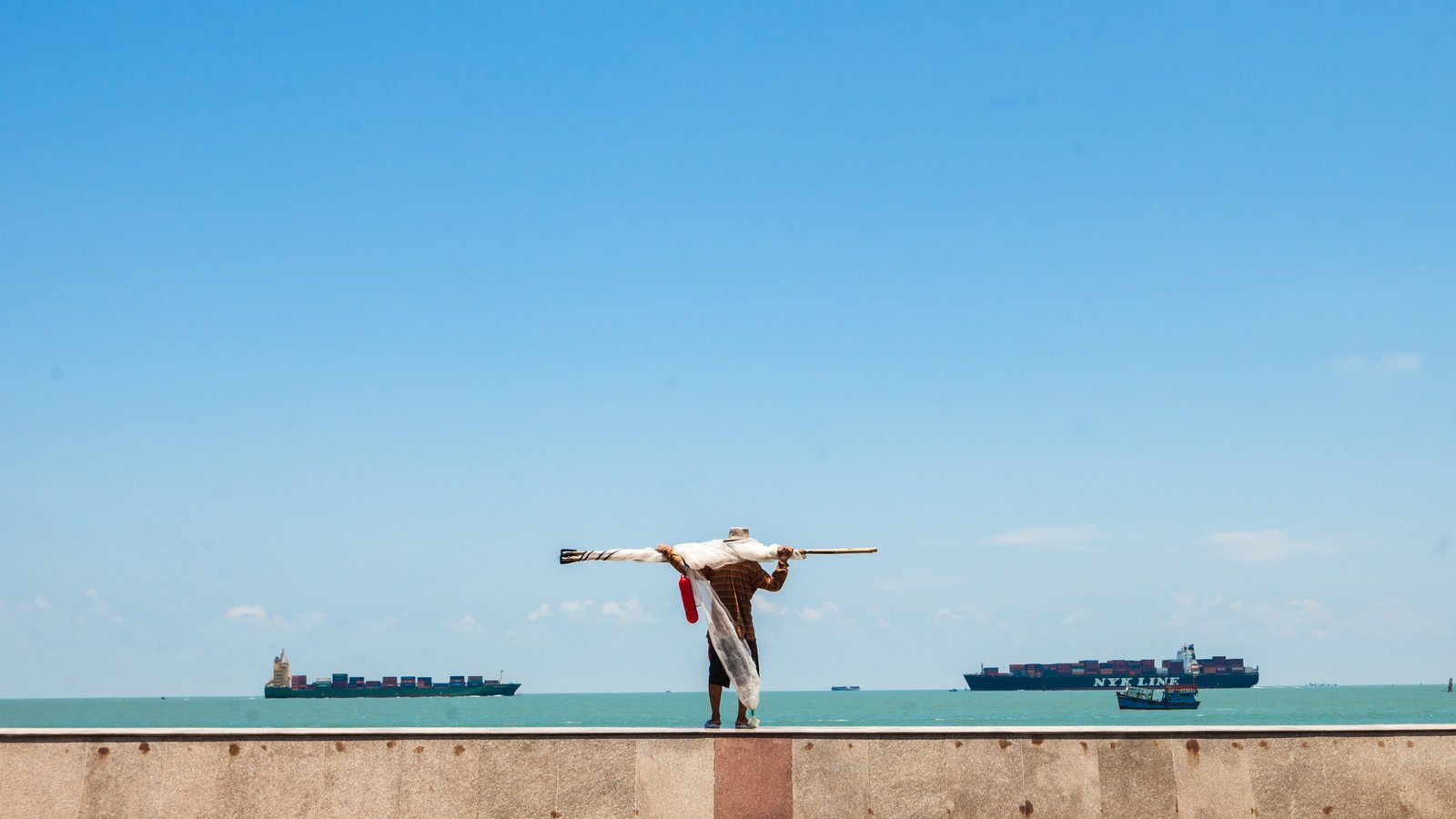 A man carrying fishing gear on a seawall in Vũng Tàu, container ships passing in the bay